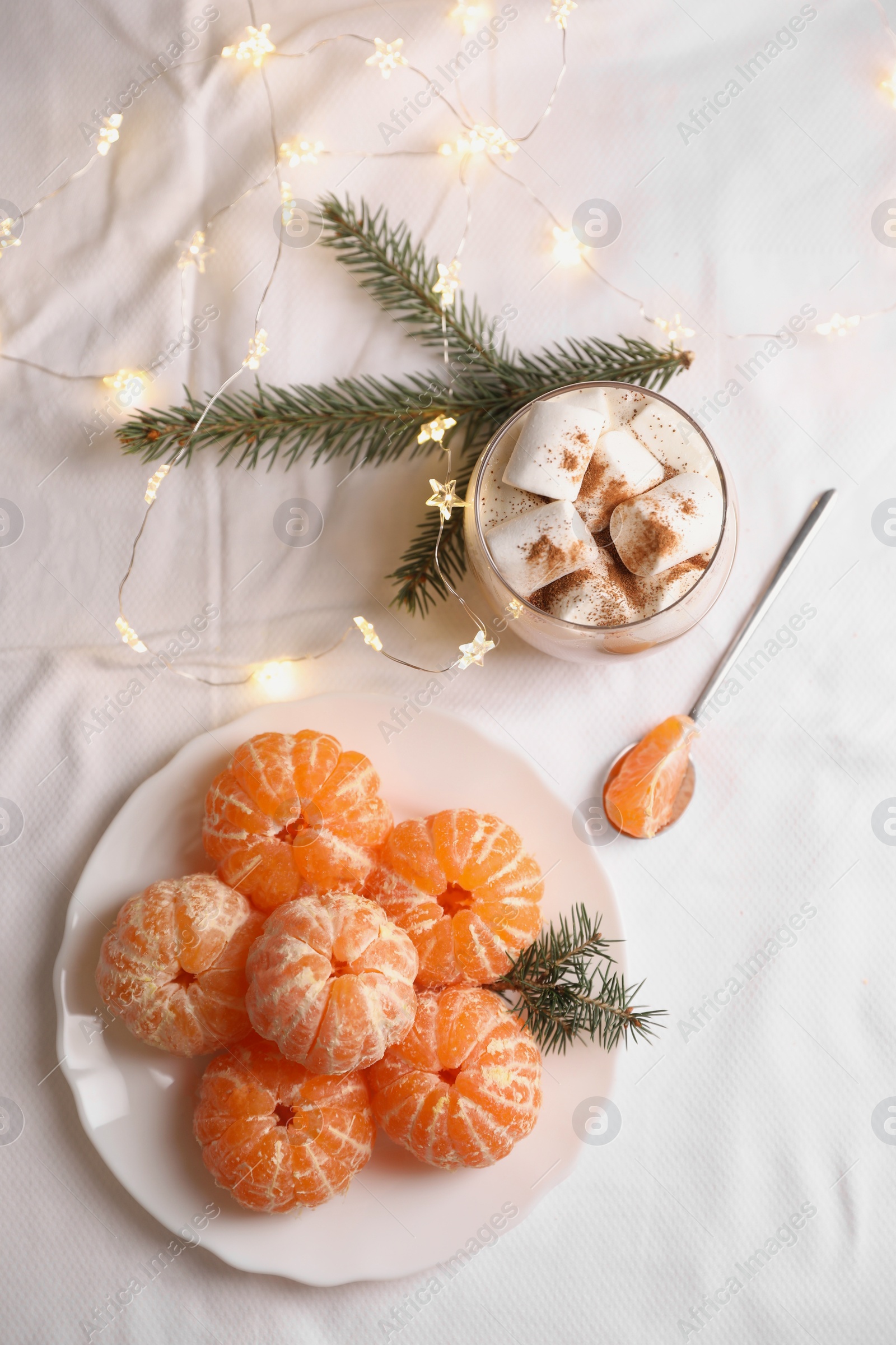 Photo of Peeled delicious ripe tangerines, festive lights and glass of drink with marshmallows on white bedsheet, flat lay