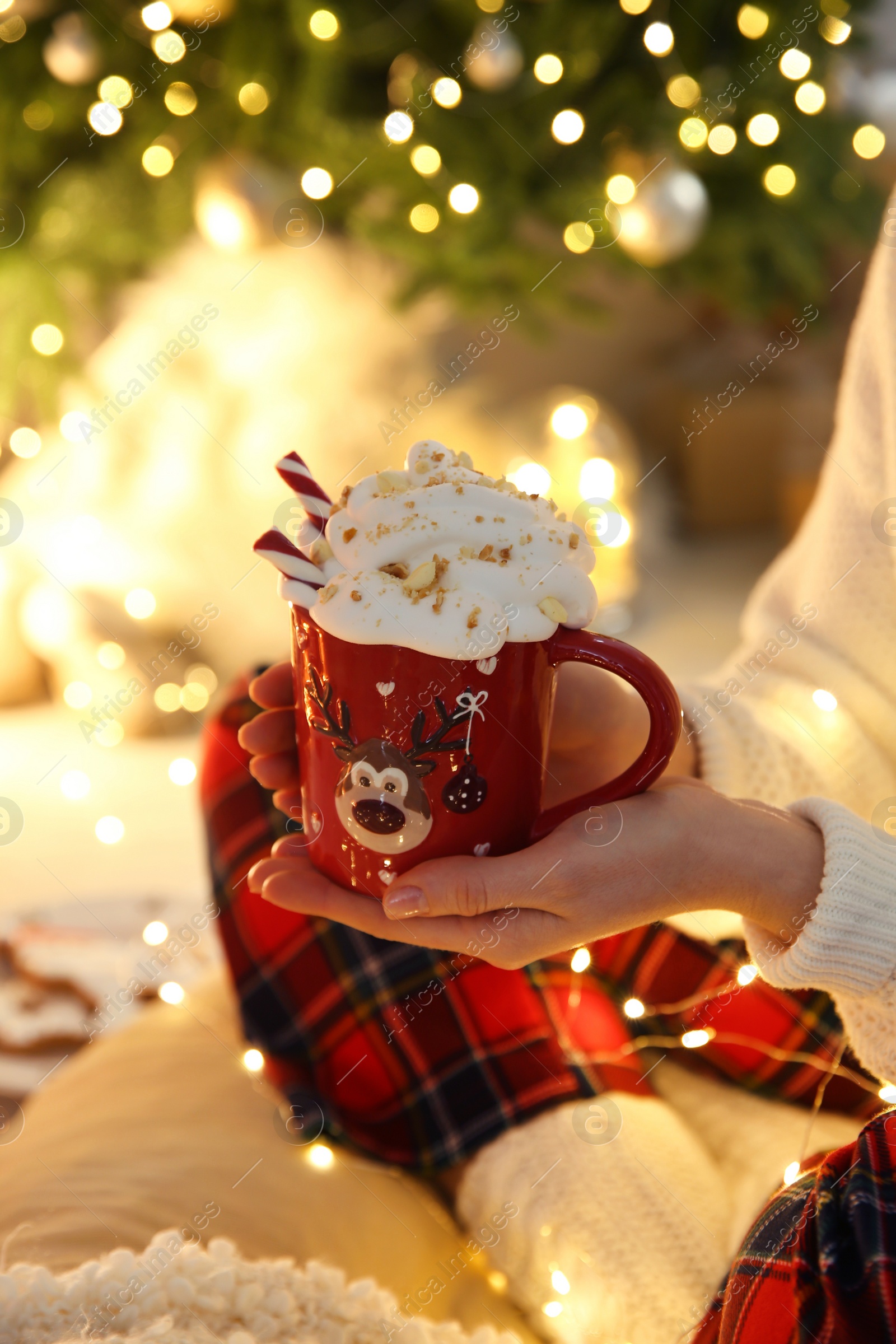 Photo of Woman holding cup of delicious drink with whipped cream near Christmas tree indoors, closeup