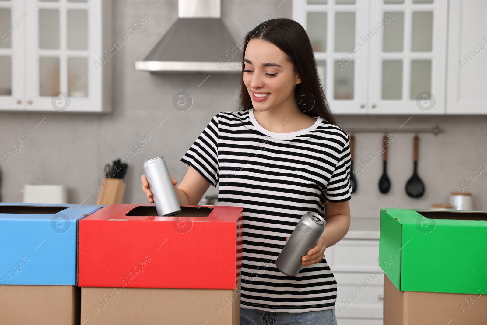 Photo of Garbage sorting. Smiling woman throwing metal can into cardboard box in kitchen