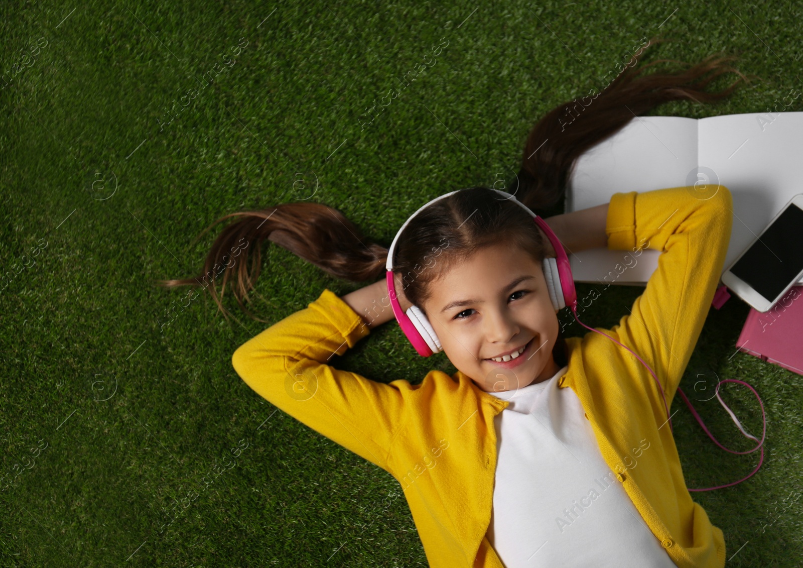 Photo of Cute little girl listening to audiobook on grass, top view