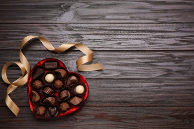 Photo of Heart shaped box with delicious chocolate candies and ribbon on wooden table, flat lay. Space for text