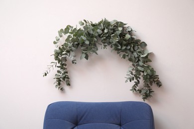 Photo of Beautiful garland made of eucalyptus branches hanging above armchair on white wall indoors