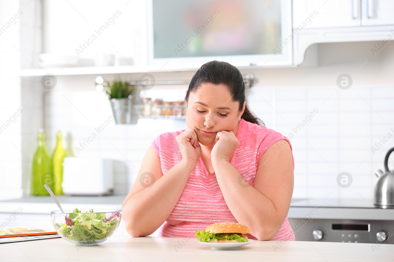 Photo of Sad overweight woman choosing between salad and burger in kitchen. Healthy diet