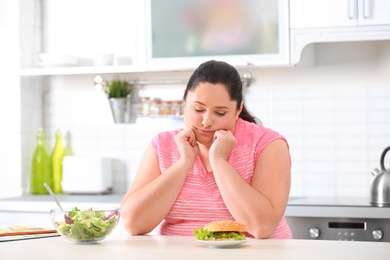 Sad overweight woman choosing between salad and burger in kitchen. Healthy diet