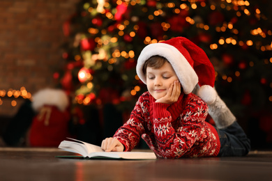 Photo of Little boy in Santa Claus cap reading book near Christmas tree at home