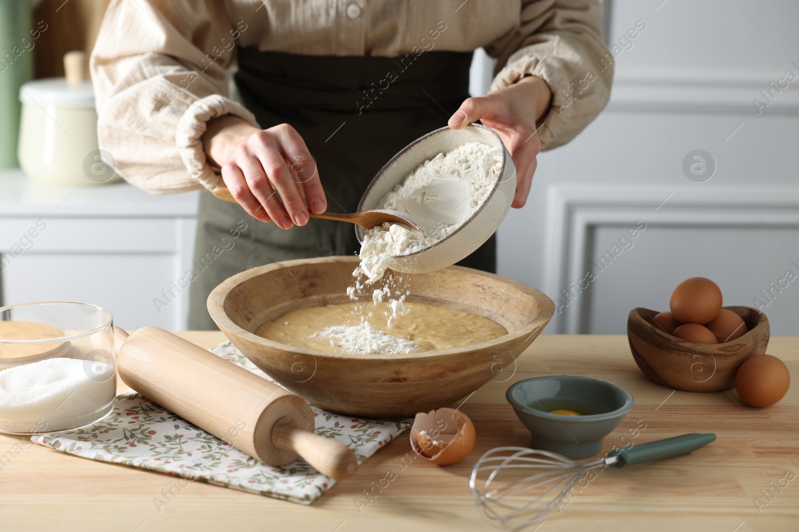 Photo of Making dough. Woman adding flour into bowl at wooden table in kitchen, closeup