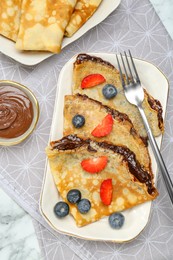 Photo of Tasty crepes with chocolate paste and berries served on white marble table, flat lay