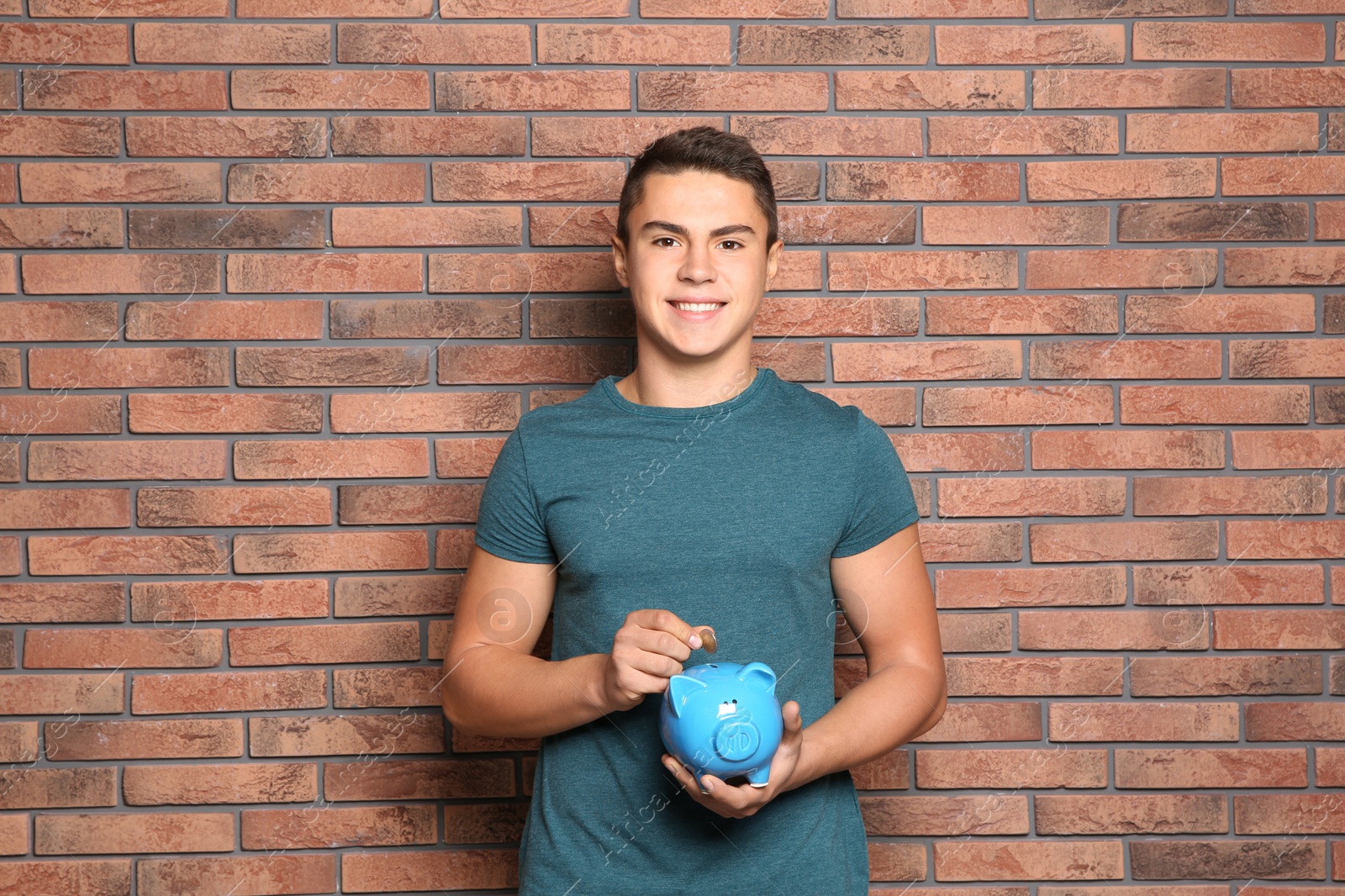 Photo of Teenage boy putting coin into piggy bank near brick wall