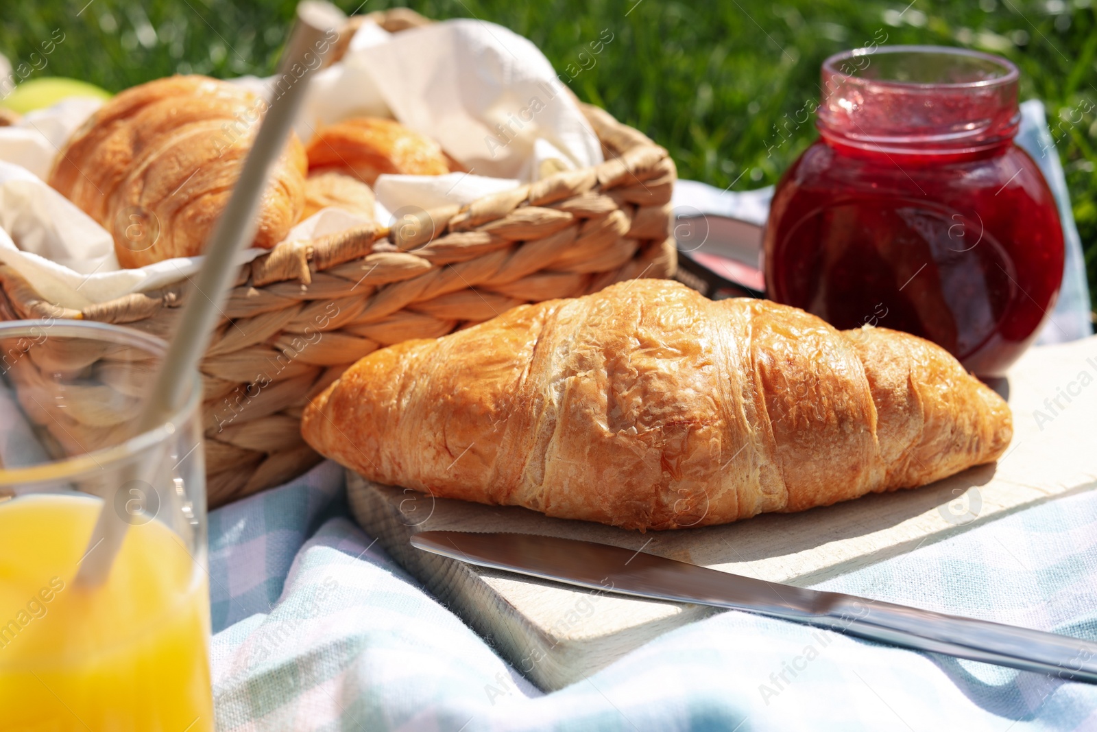 Photo of Blanket with juice, jam and croissants for picnic on green grass, closeup