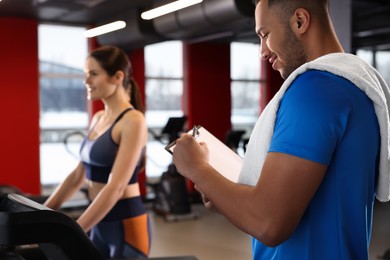 Photo of Happy trainer writing down workout plan while woman doing exercise in modern gym