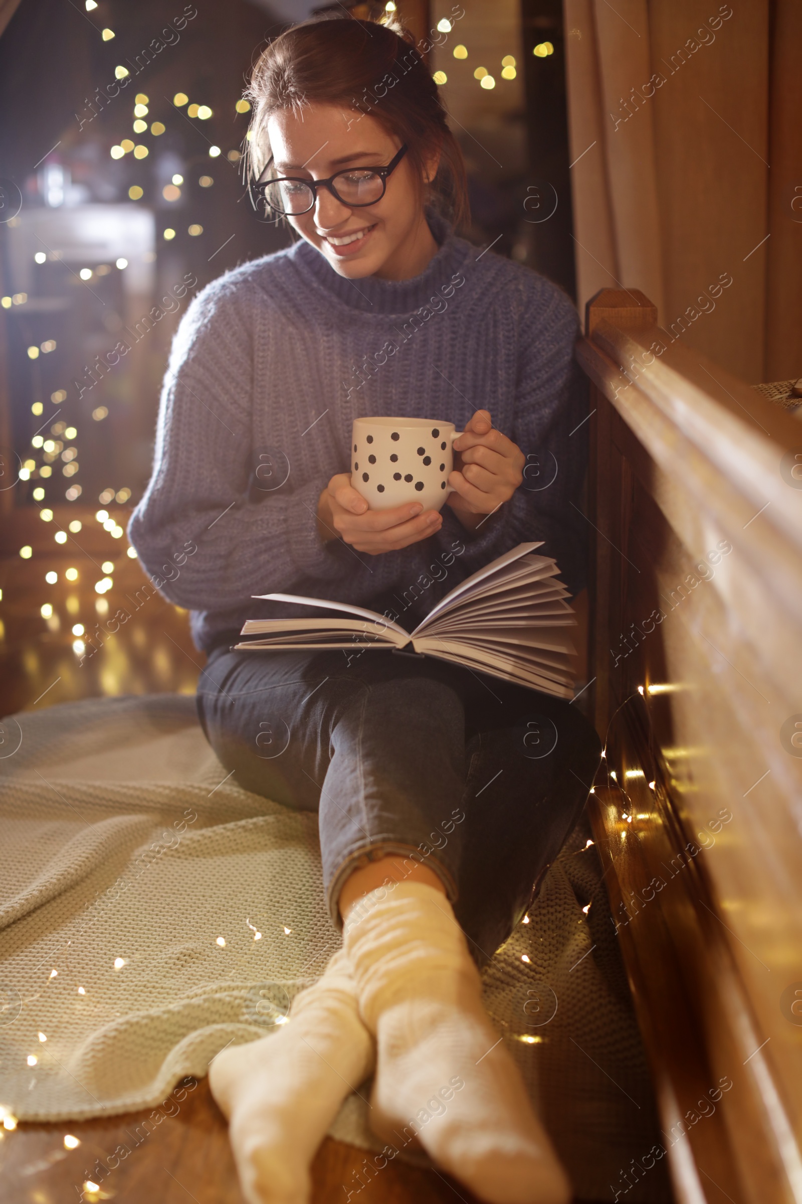 Photo of Woman with cup of hot beverage reading book at home in winter evening