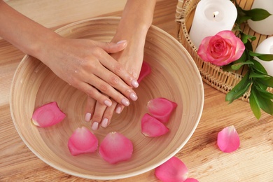 Photo of Woman soaking her hands in bowl with water and petals on wooden table, top view. Spa treatment
