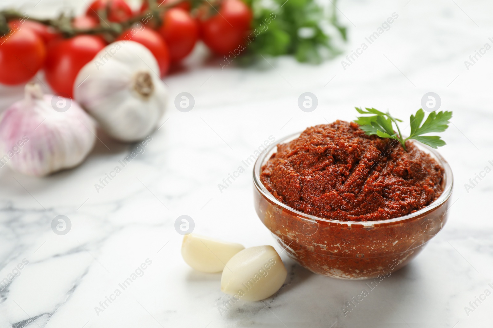 Photo of Delicious adjika sauce in glass bowl and ingredients on white marble table, closeup