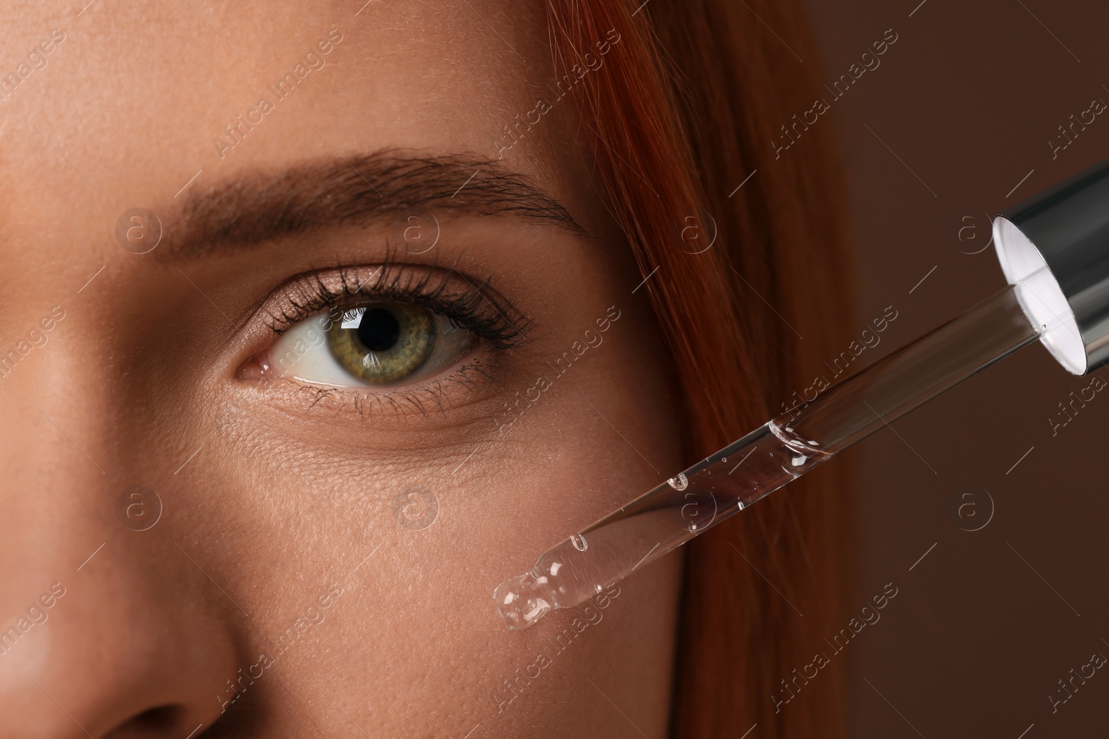 Photo of Beautiful young woman applying cosmetic serum onto her face on brown background, closeup