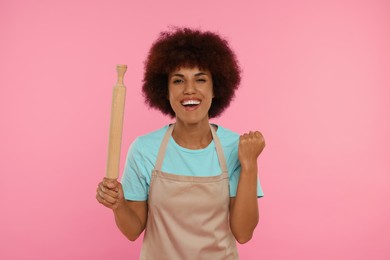 Photo of Happy young woman in apron holding rolling pin on pink background