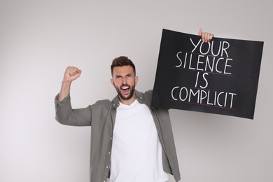 Photo of Emotional man holding sign with phrase Your Silence Is Complicit on light background. End racism