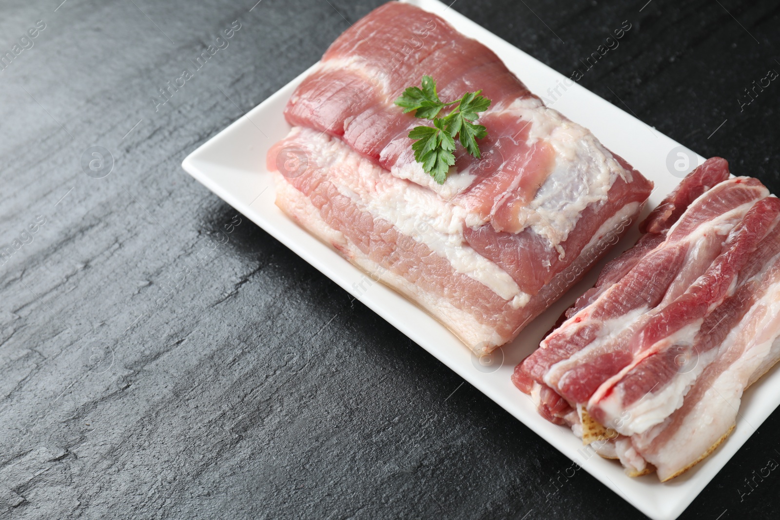 Photo of Pieces of raw pork belly and parsley on black textured table, above view. Space for text