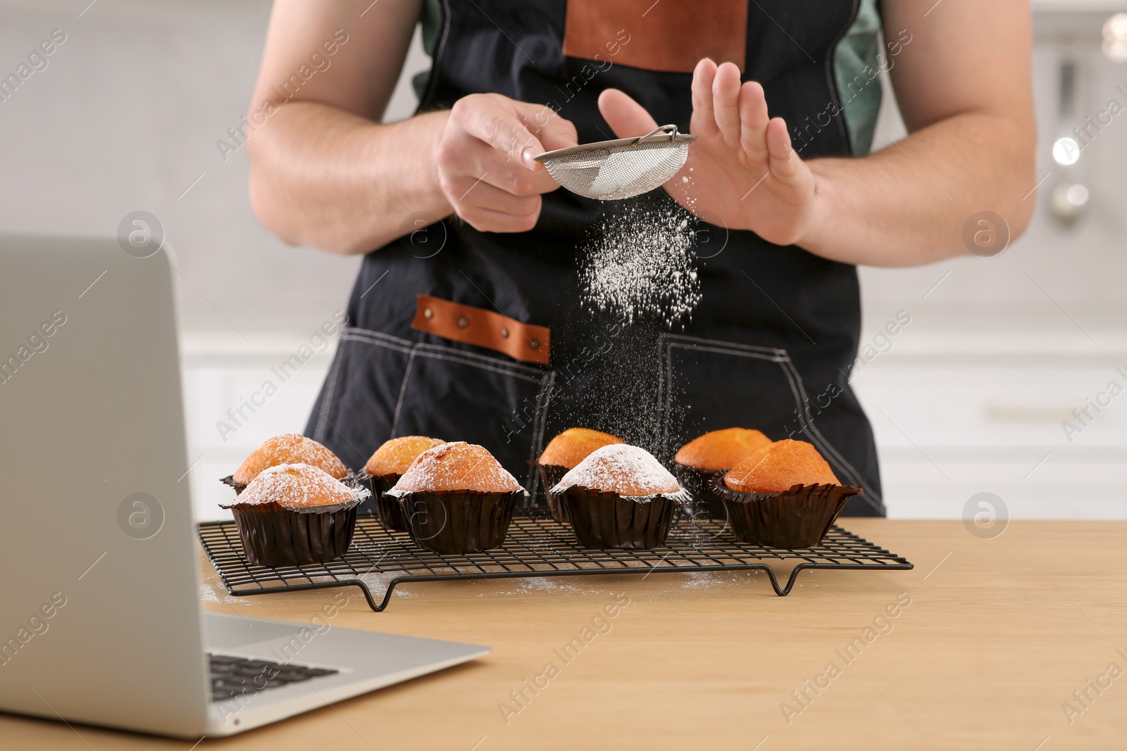 Photo of Man decorating muffins with powdered sugar while watching online cooking course via laptop in kitchen, closeup. Time for hobby