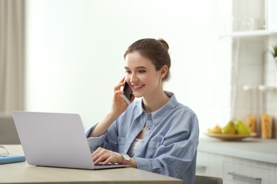 Photo of Young woman talking on phone while using laptop at table indoors