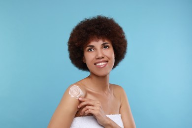 Beautiful young woman applying body cream onto shoulder on light blue background