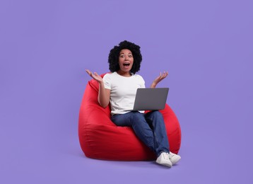 Emotional young woman with laptop sitting on beanbag chair against purple background