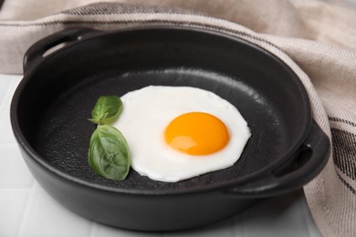 Tasty fried egg with basil in pan on white tiled table, closeup