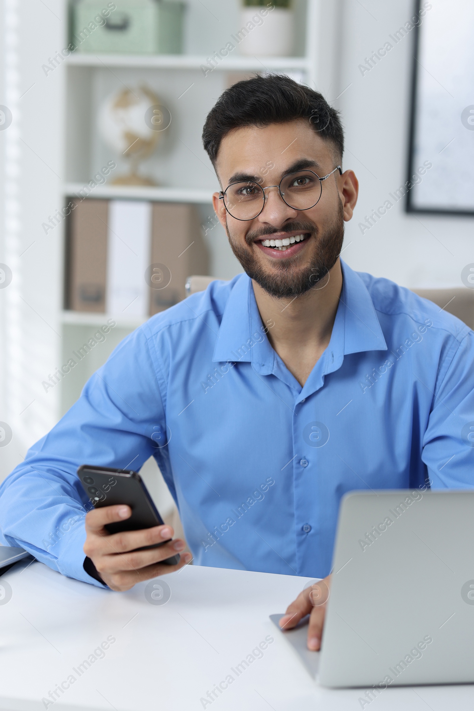 Photo of Happy young man using smartphone while working with laptop at white table in office