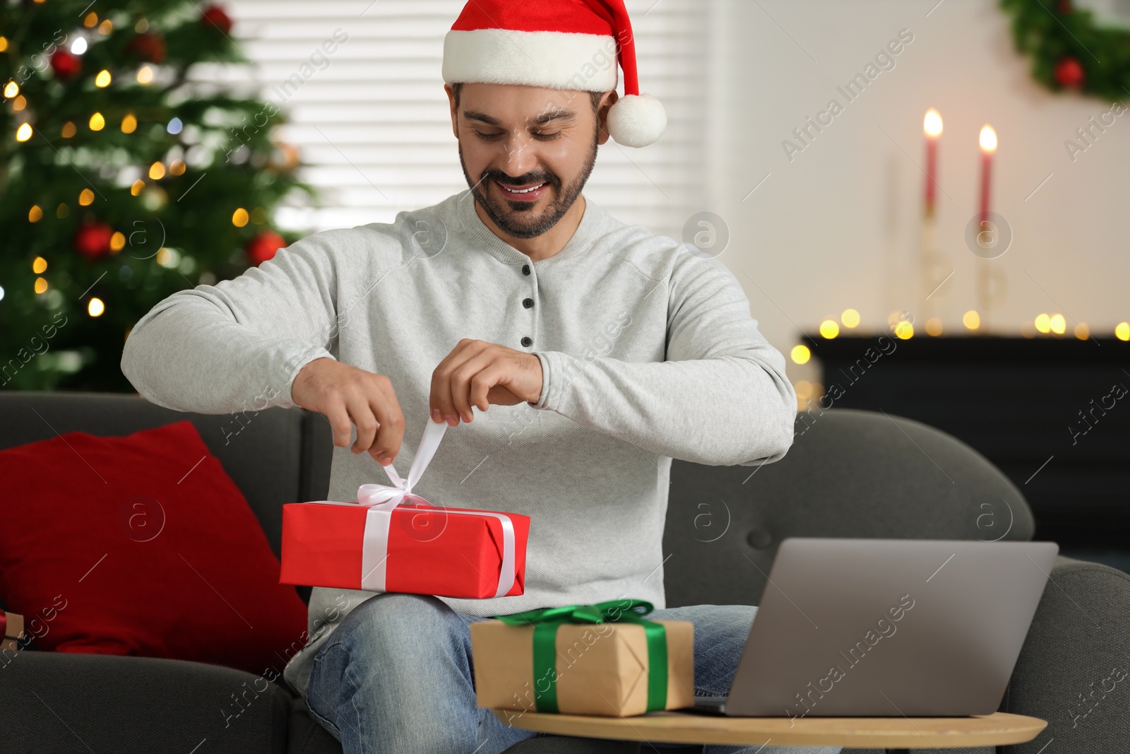 Photo of Celebrating Christmas online with exchanged by mail presents. Happy man in Santa hat opening gift box during video call on laptop at home