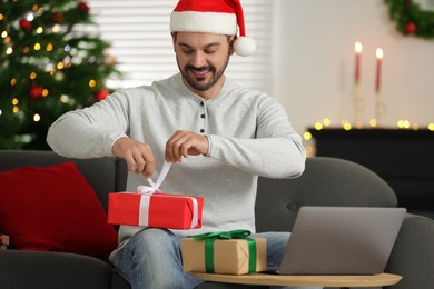 Photo of Celebrating Christmas online with exchanged by mail presents. Happy man in Santa hat opening gift box during video call on laptop at home