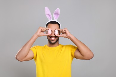 Happy African American man in bunny ears headband covering eyes with Easter eggs on gray background