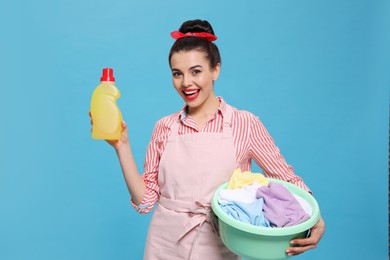 Photo of Housewife holding bottle of cleaning product and basin with clothes on light blue background