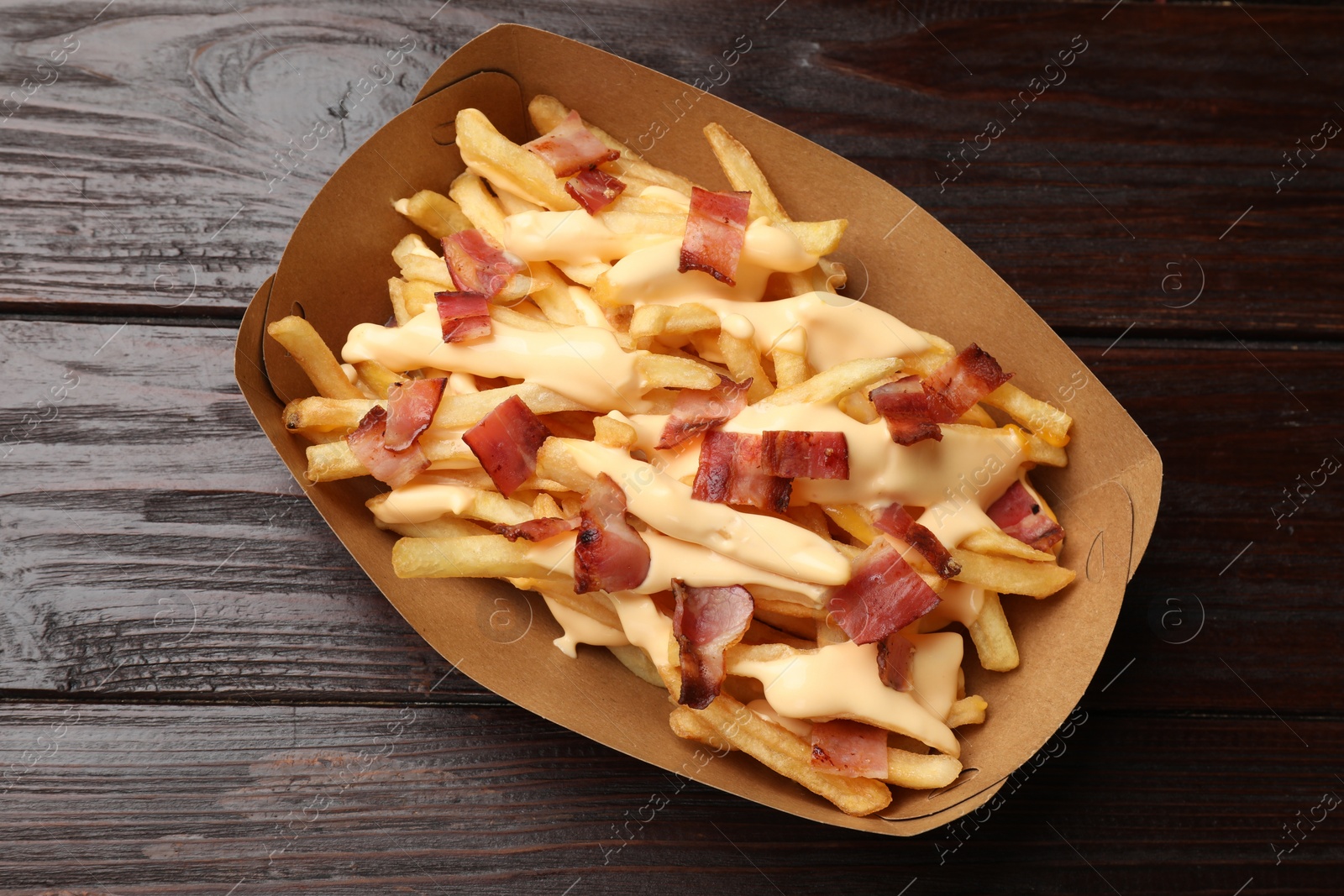 Photo of Tasty potato fries, cheese sauce and bacon in paper container on wooden table, top view