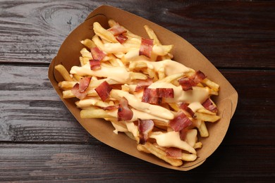 Tasty potato fries, cheese sauce and bacon in paper container on wooden table, top view