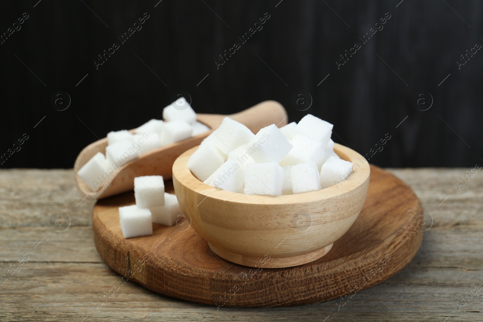Photo of White sugar cubes in bowl and scoop on wooden table against dark background, closeup