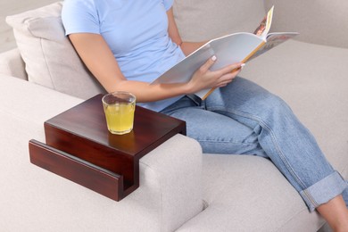 Photo of Glass of juice on wooden sofa armrest table. Woman reading magazine at home, closeup