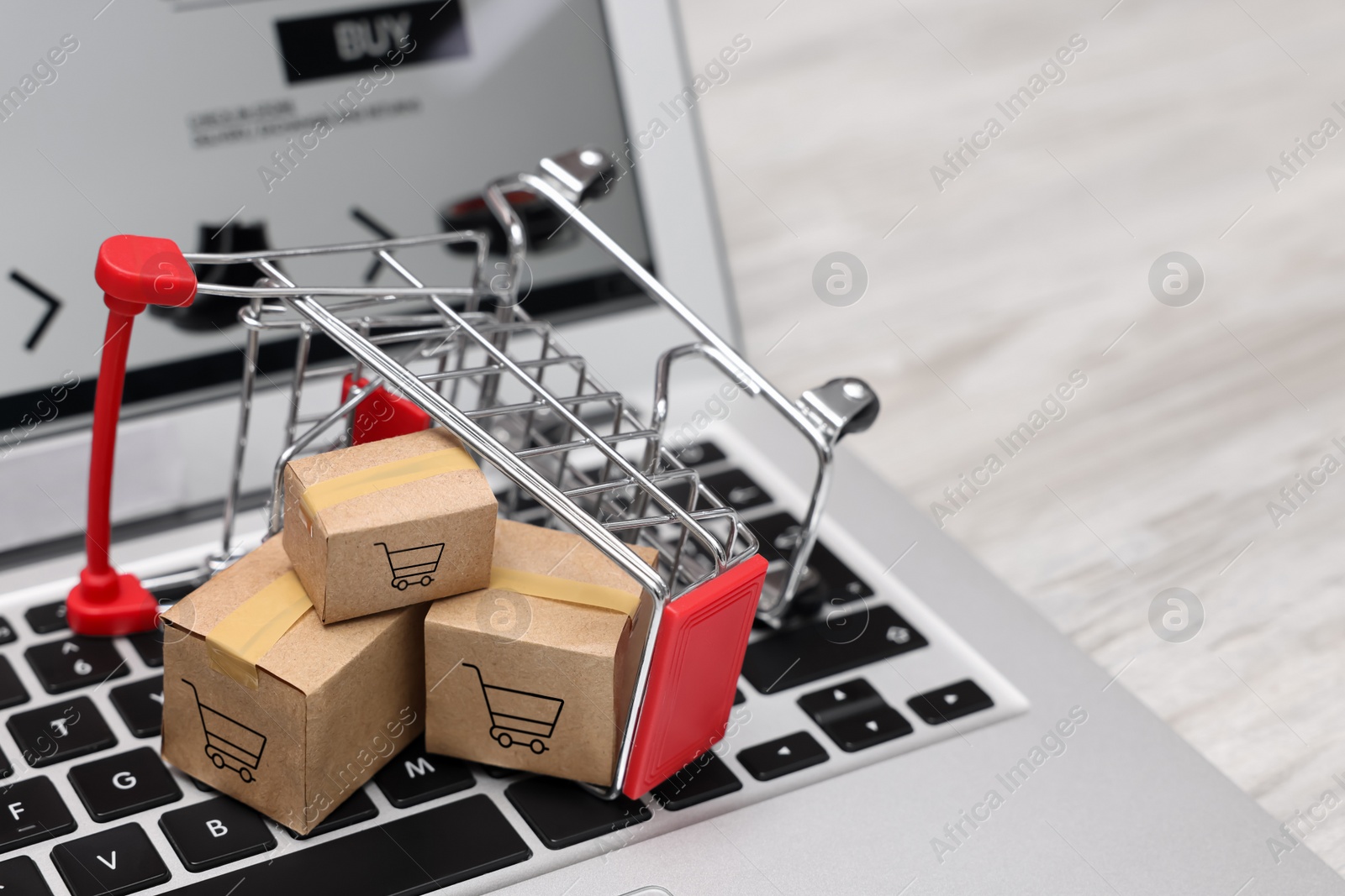 Image of Internet store. Small cardboard boxes, shopping cart and laptop on light wooden table, closeup