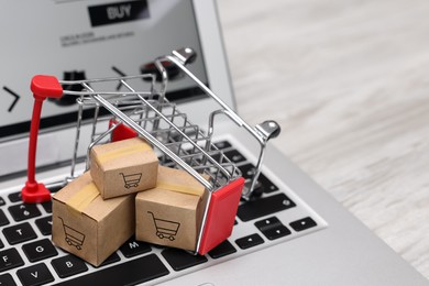 Internet store. Small cardboard boxes, shopping cart and laptop on light wooden table, closeup