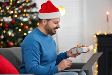 Celebrating Christmas online with exchanged by mail presents. Happy man in Santa hat with gift box during video call on laptop at home