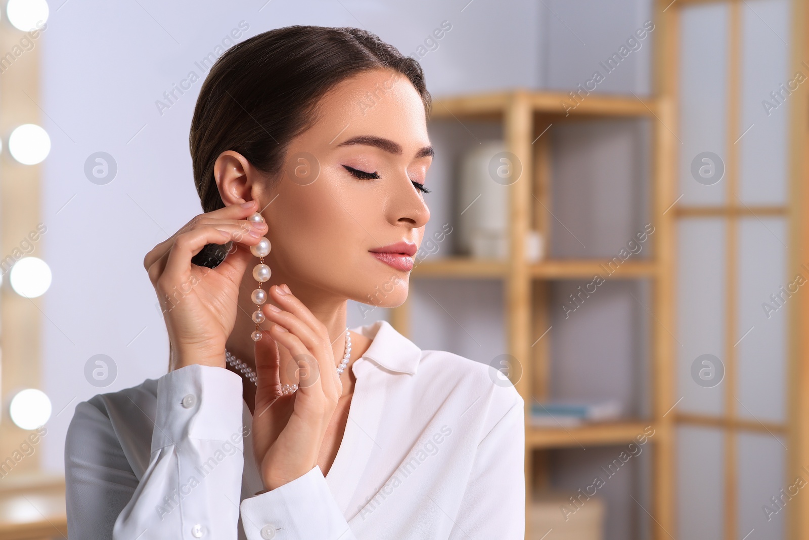 Photo of Young woman trying on elegant pearl earring indoors, space for text