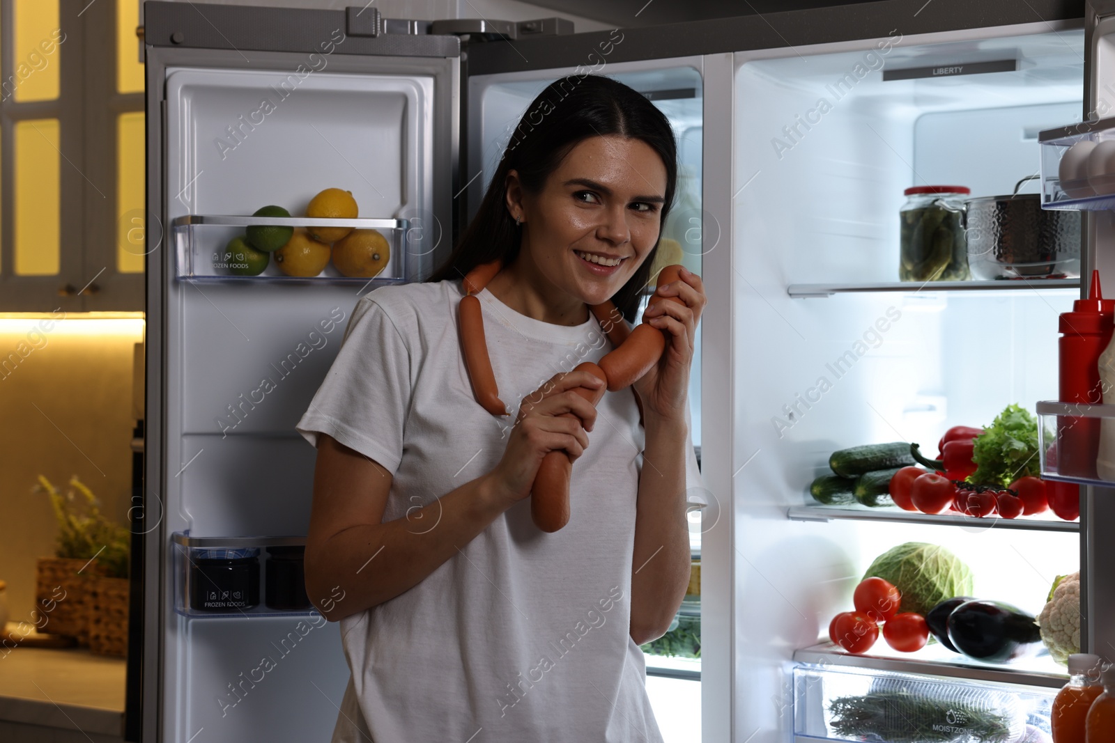 Photo of Young woman with sausages near modern refrigerator in kitchen at night