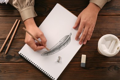 Woman drawing feather with graphite pencil in sketchbook at wooden table, top view