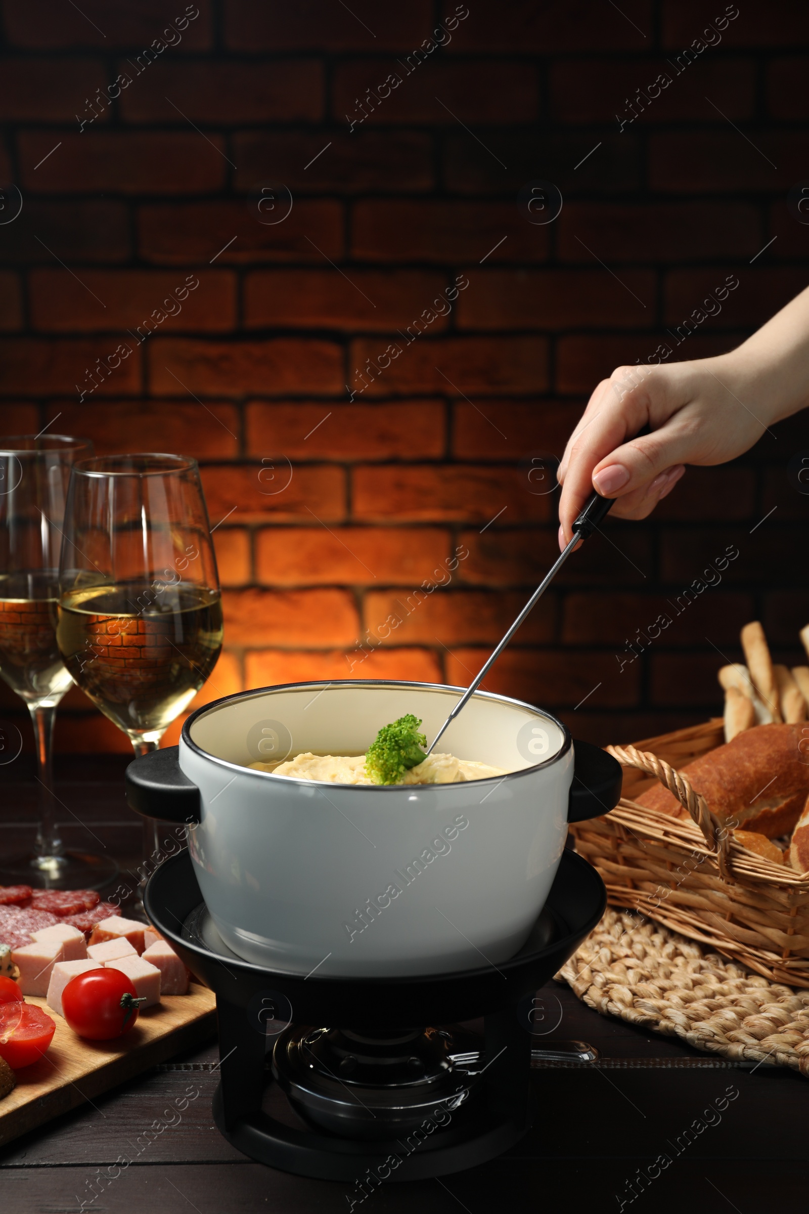 Photo of Woman dipping piece of broccoli into fondue pot with melted cheese at wooden table with wine and snacks, closeup