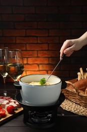 Photo of Woman dipping piece of broccoli into fondue pot with melted cheese at wooden table with wine and snacks, closeup