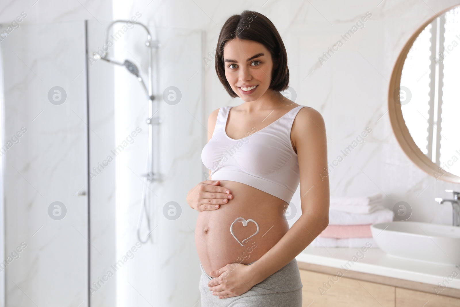 Photo of Young pregnant woman with cosmetic product on belly in bathroom