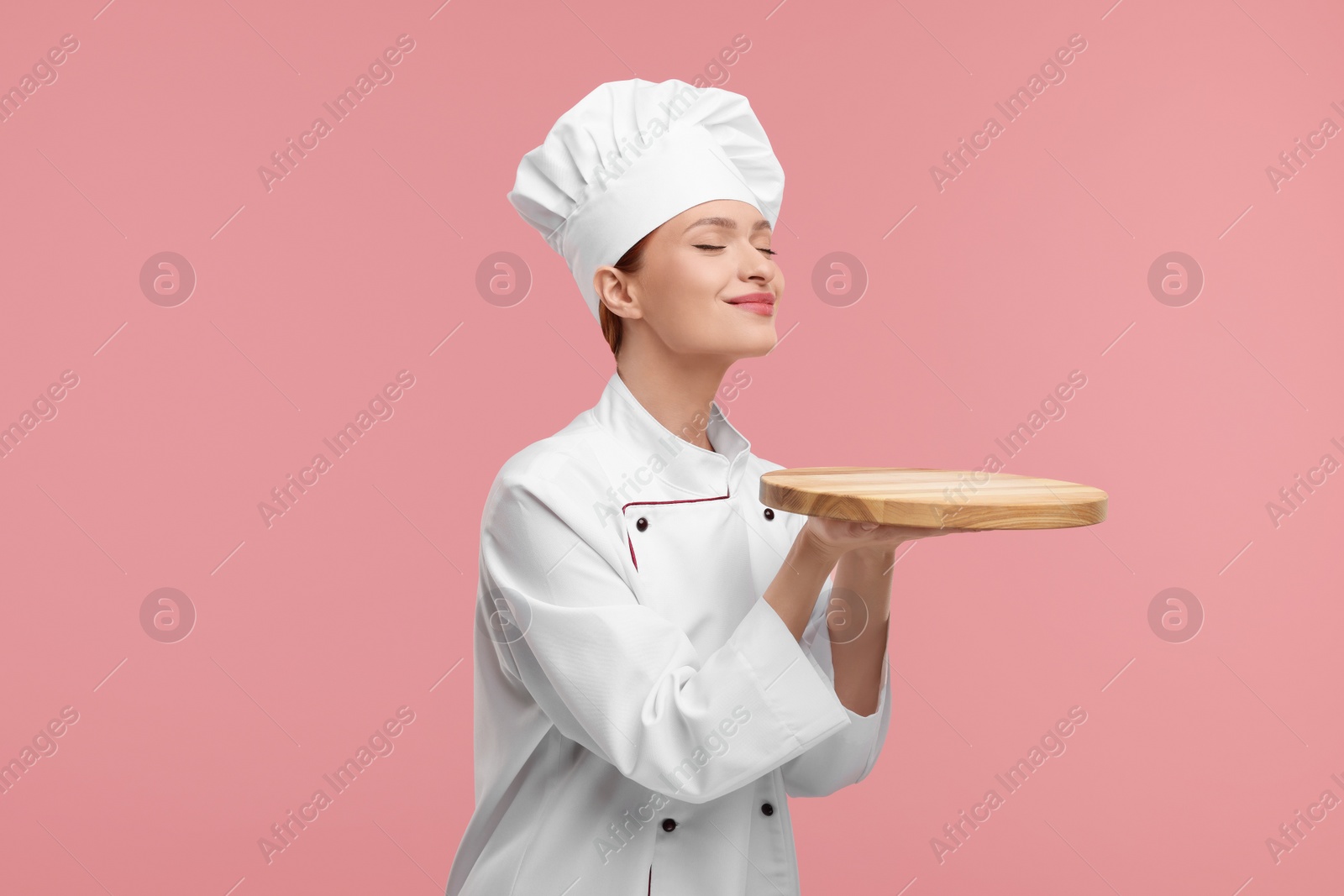 Photo of Young chef in uniform holding empty wooden board on pink background