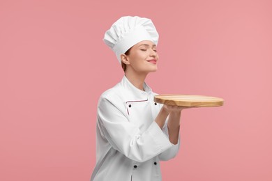 Photo of Young chef in uniform holding empty wooden board on pink background