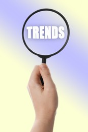 Image of Searching new and popular trends. Woman holding magnifying glass over word on color background, closeup