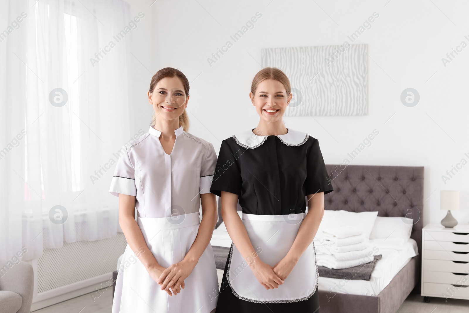 Photo of Professional chambermaids in uniform standing near bed indoors