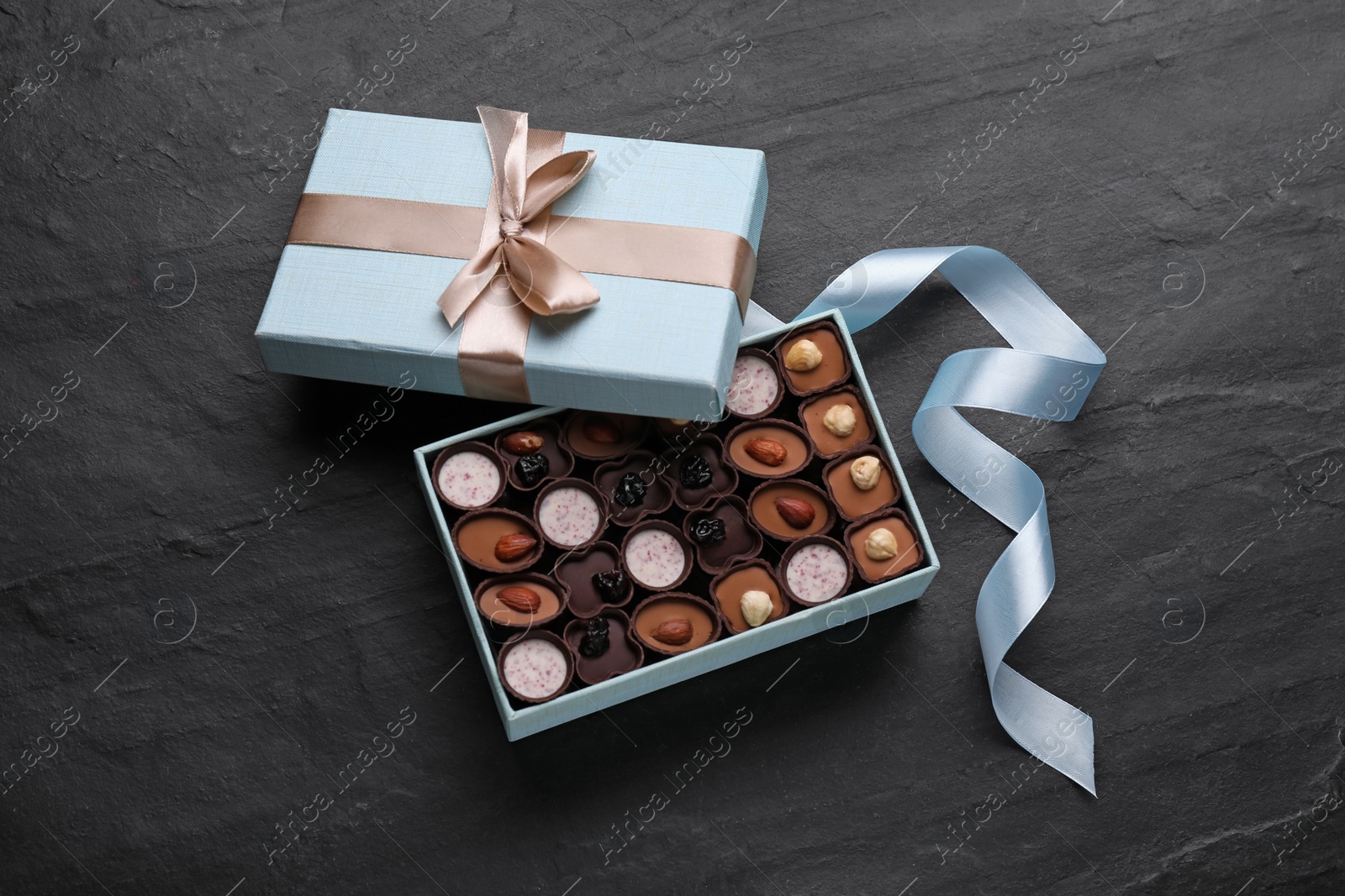 Photo of Open box of delicious chocolate candies and light blue ribbon on black table, flat lay