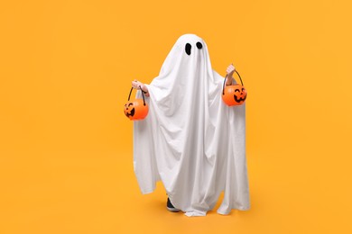 Photo of Child in white ghost costume holding pumpkin buckets on orange background, space for text. Halloween celebration